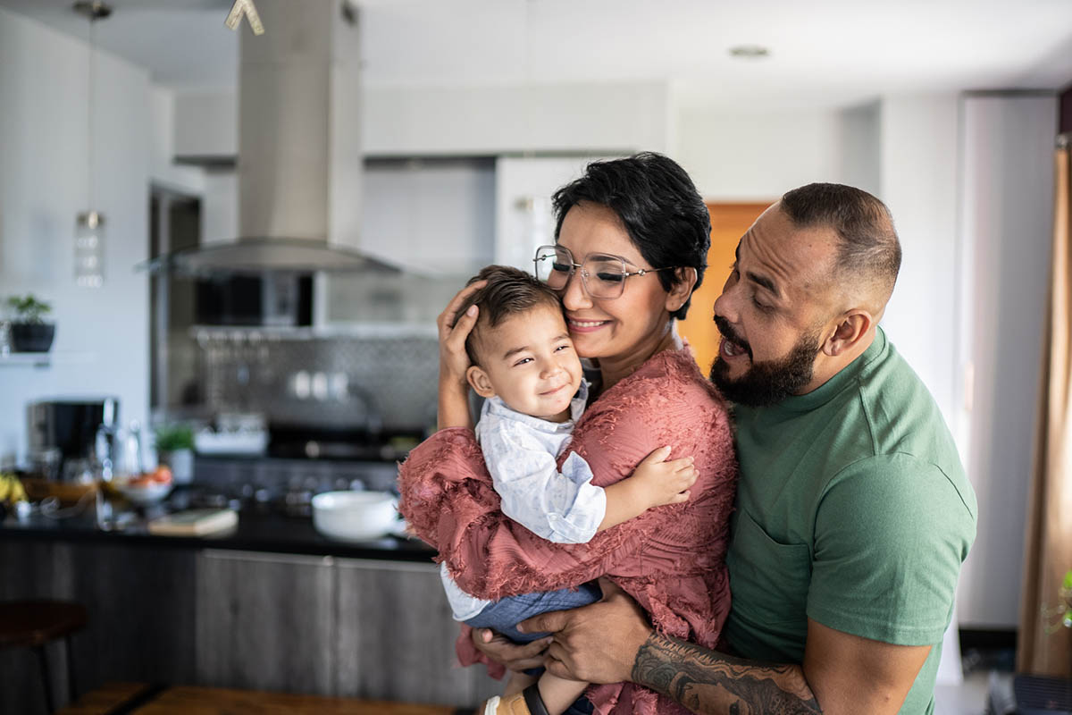 A mother holds her young child, smiling, while the dad makes a funny face