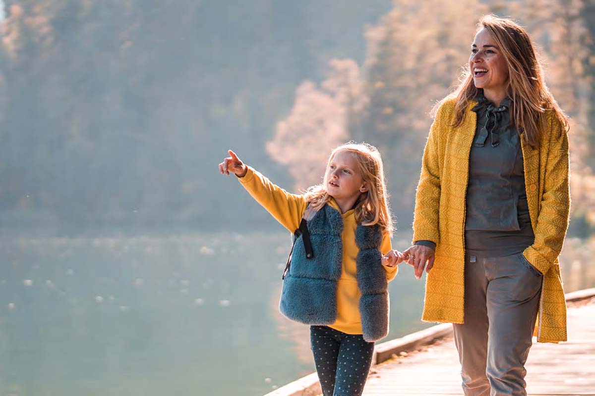 A woman and girl walk on a path by a lake on a fall day. The girl is pointing up at something.