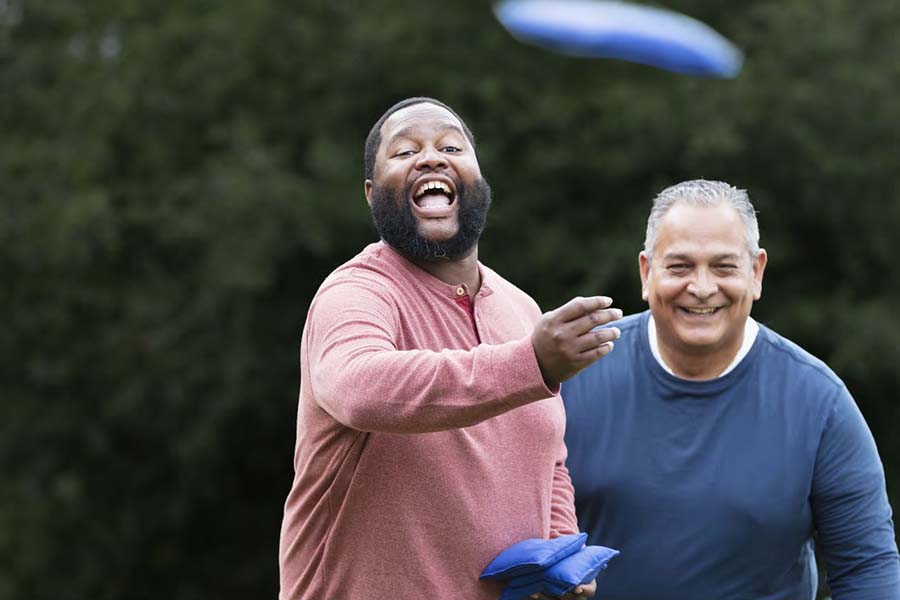 Two men smile outdoors, as one tosses a beanbag