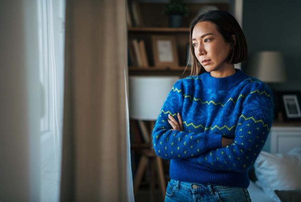 A young woman stands by the window, depressed, her arms crossed.