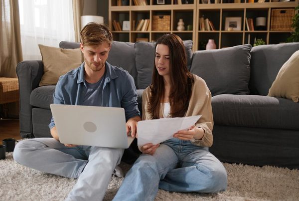 Young couple sitting on floor in front of a couch, shopping for health insurance on a laptop computer.